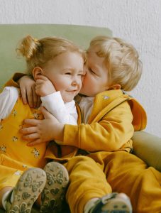 Two toddlers dressed in yellow and white. The young body platonically and affectionately kisses the young girl (perhaps his sister)